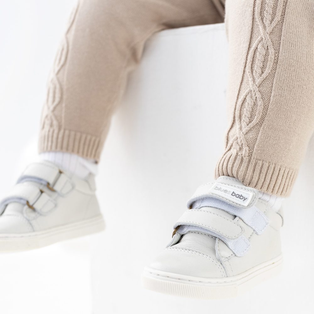 A pair of child's legs wearing knitted beige leggings and white trainers by Blues Baby