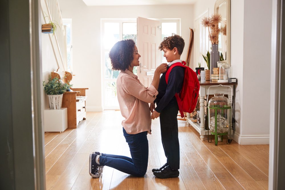 A woman knelt in the hallway of a house straightening the tie of a young boy in school uniform wearing a red backpack 