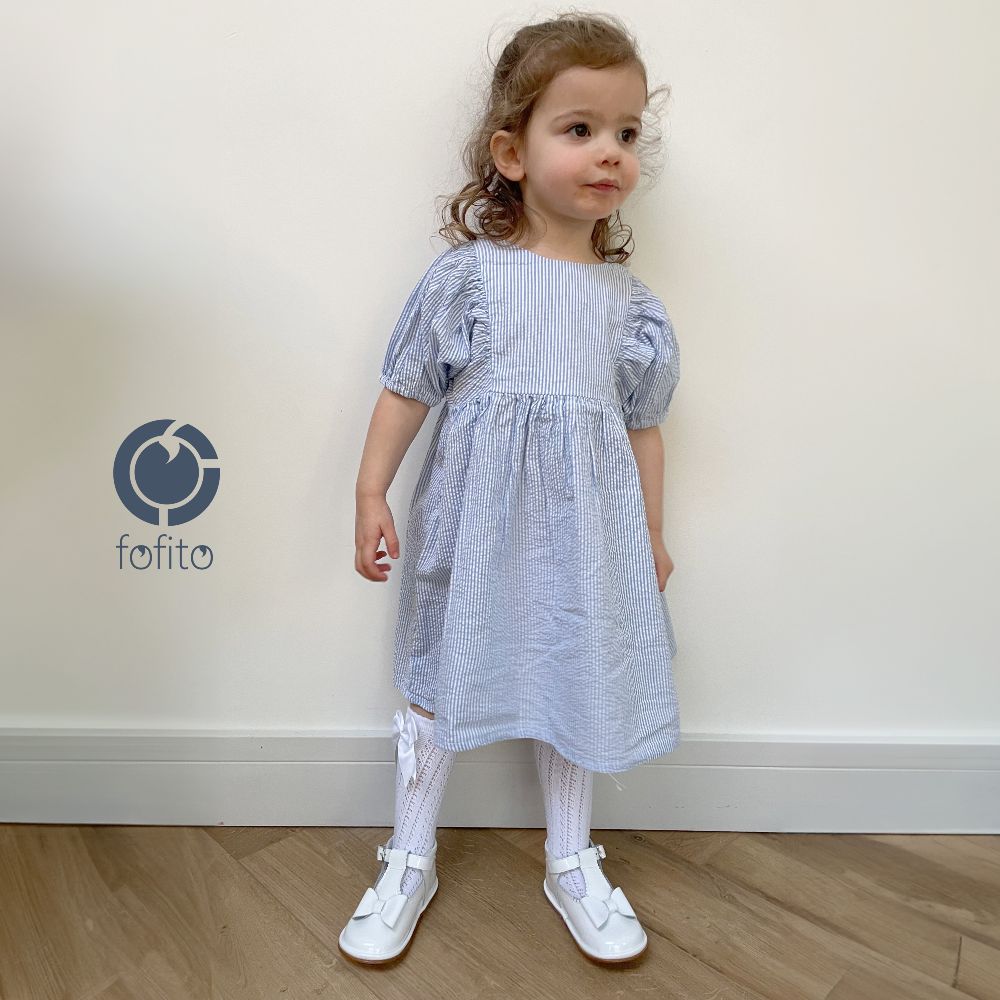 A young girl stood on a room wearing a blue dress and white patent bow shoes 