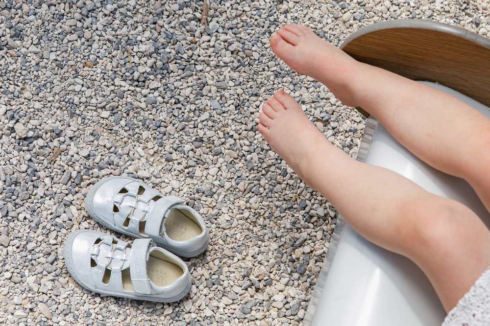 A picture of a young child's bare feet with a pair of Froddo sandals on the gravel floor