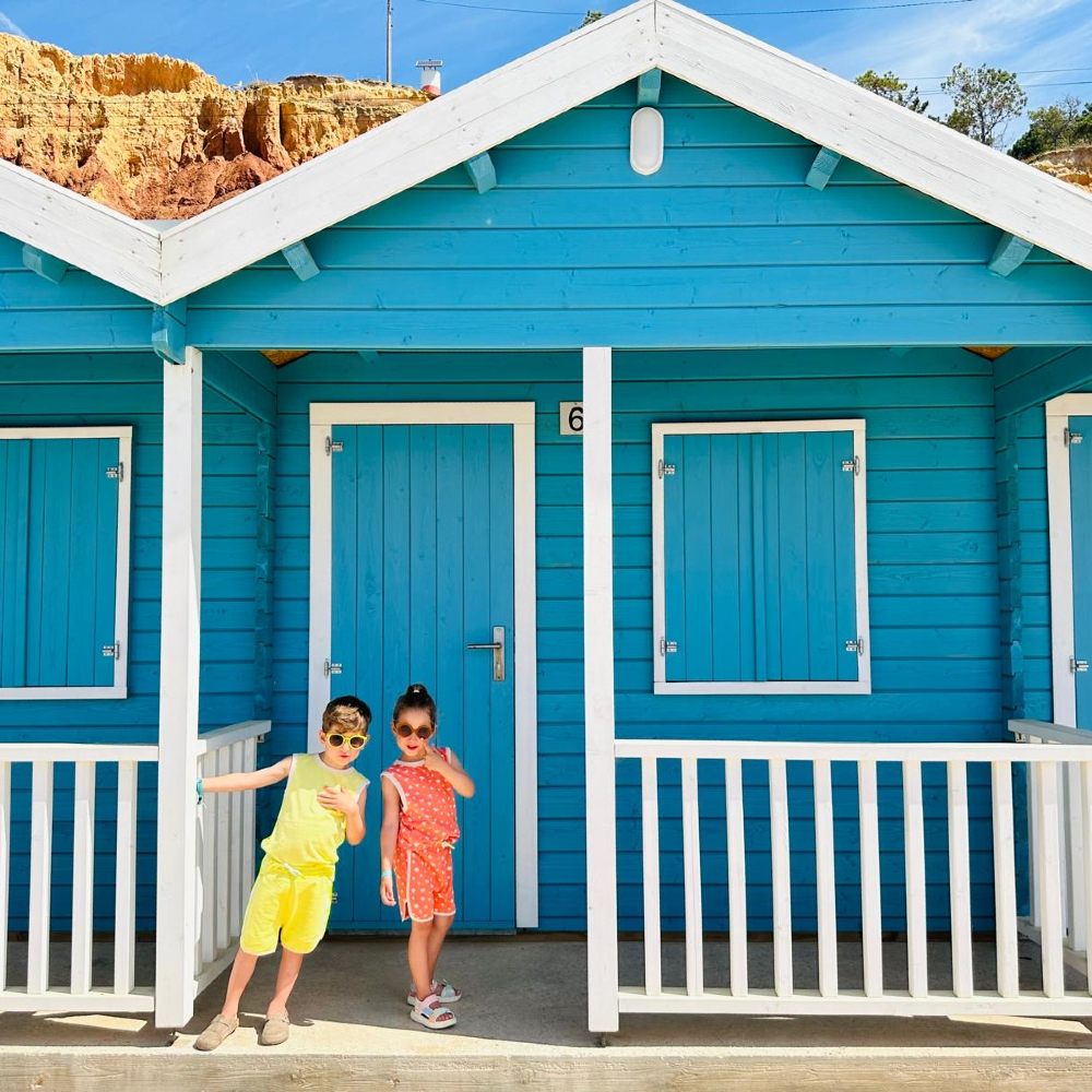 Two young children in shorts and T-shirt sets stood outside a blue and white beach hut 