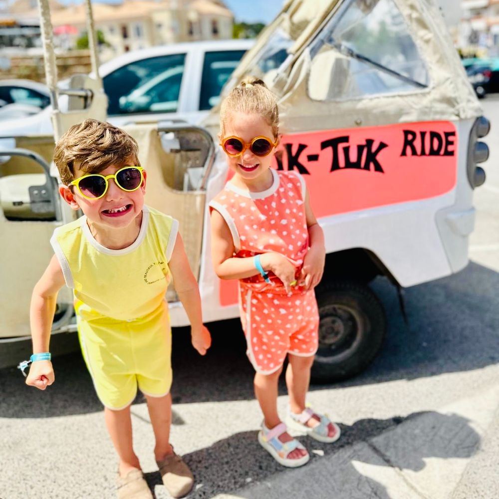 Two children stood on a pavement beside a tuk-tuk wearing shorts and T-shirts and sunglasses 