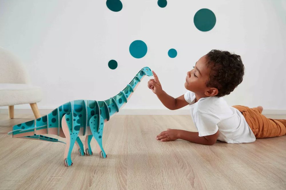 A young boy lying on the floor playing with a blue dinosaur toy by Janod