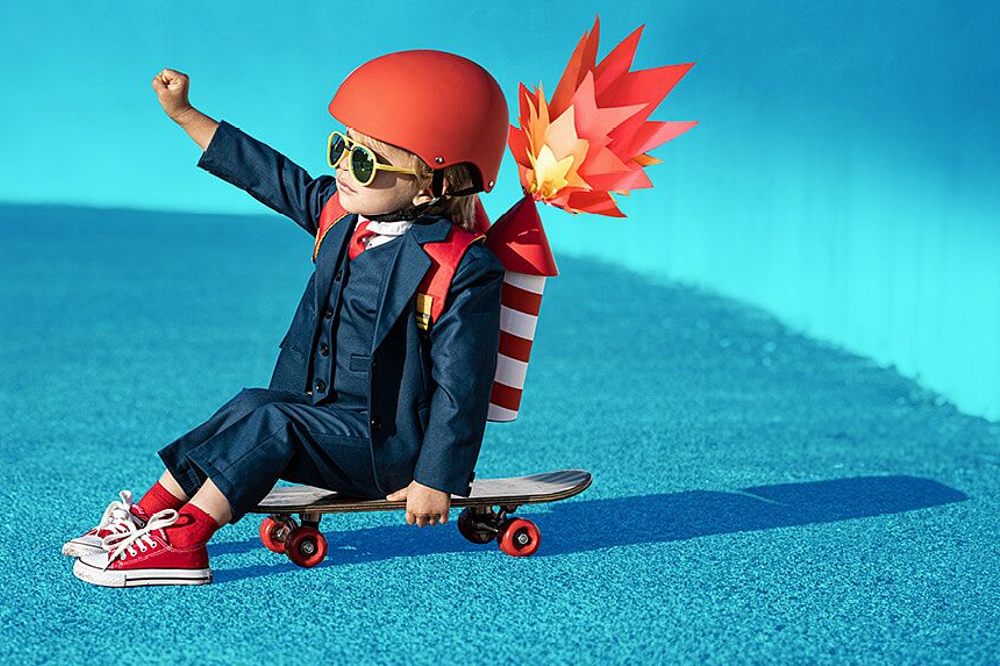 A young boy in a suit and red helmet sitting on a skateboard with a pretend rocket on his back
