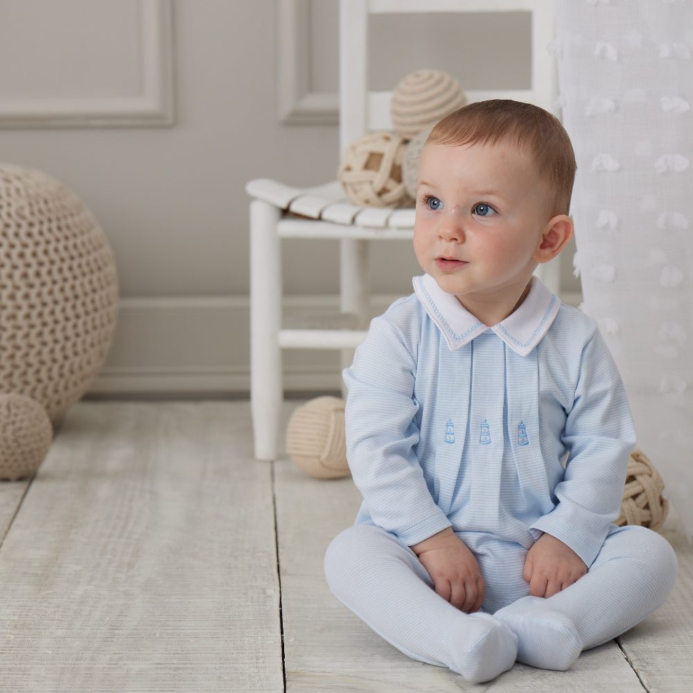 A young baby sat in a room on a white wooden floor wearing a pale blue collared babygro 