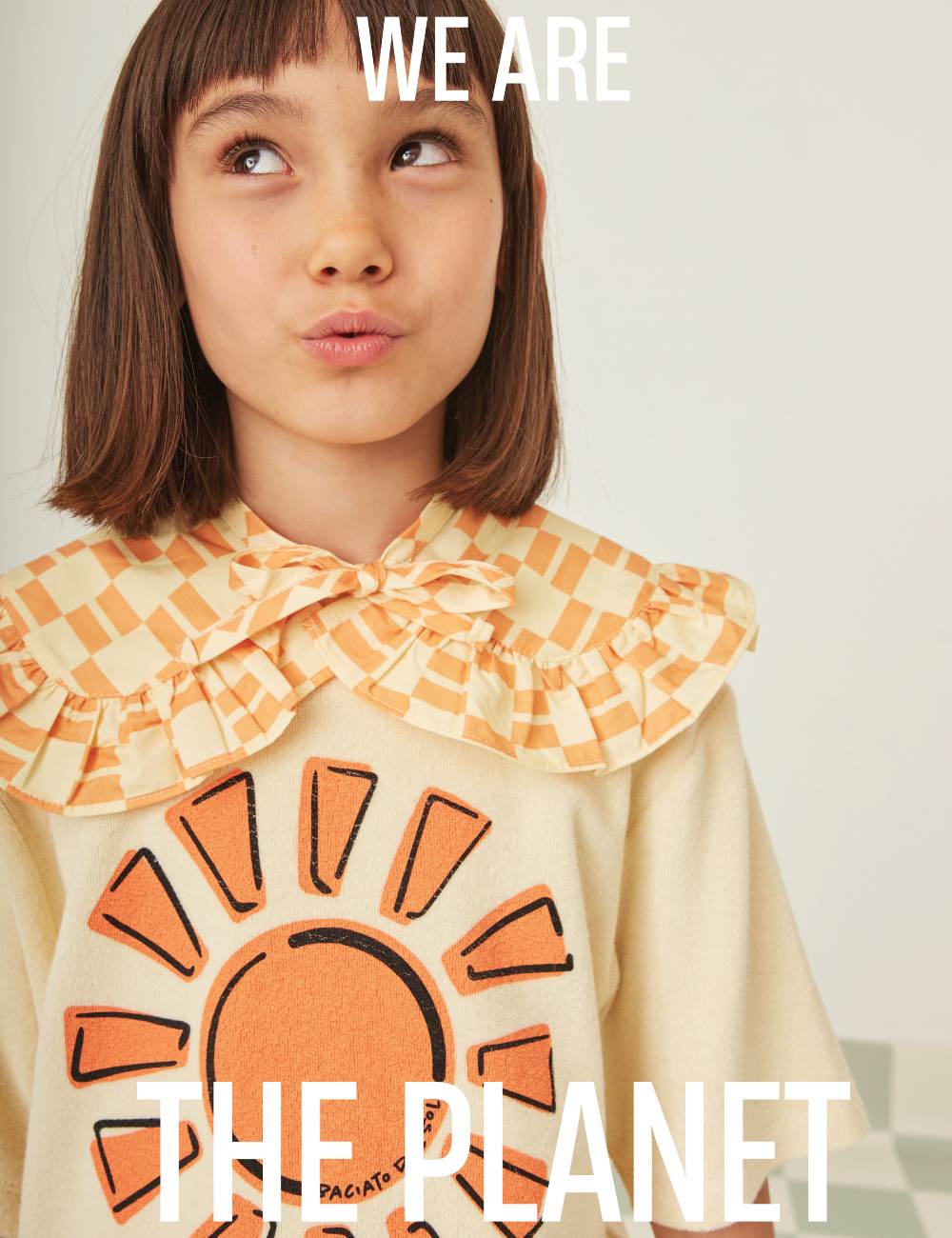 A young girl with dark hair looking upwards wearing a collared yellow top with a sun on the front with the words We Are The Planet over the image 