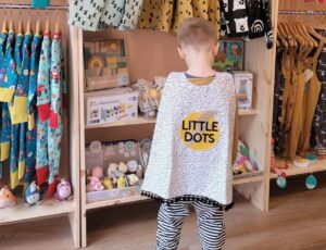 A child stood in a childrenswear shop wearing a spotted cape with Little Dots written on it