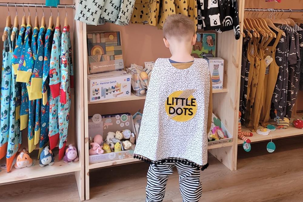 A child stood in a childrenswear shop wearing a spotted cape with Little Dots written on it