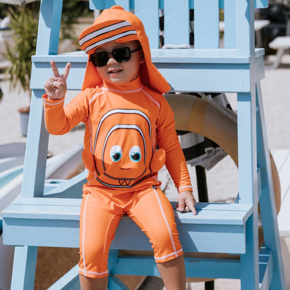 A young child sat outside on blue wooden chair wearing an orange sunsuit, hat and sunglasses 