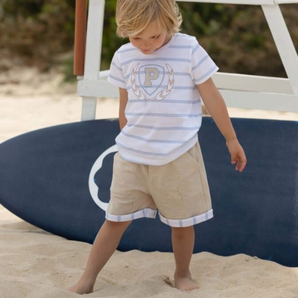A young boy on a beach beside a surfboard looking down at his feet in the sand 
