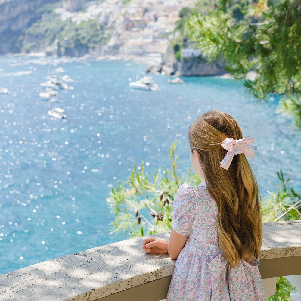 A young girl in a floral dress and hair bow with her back to the camera looking out to sea 