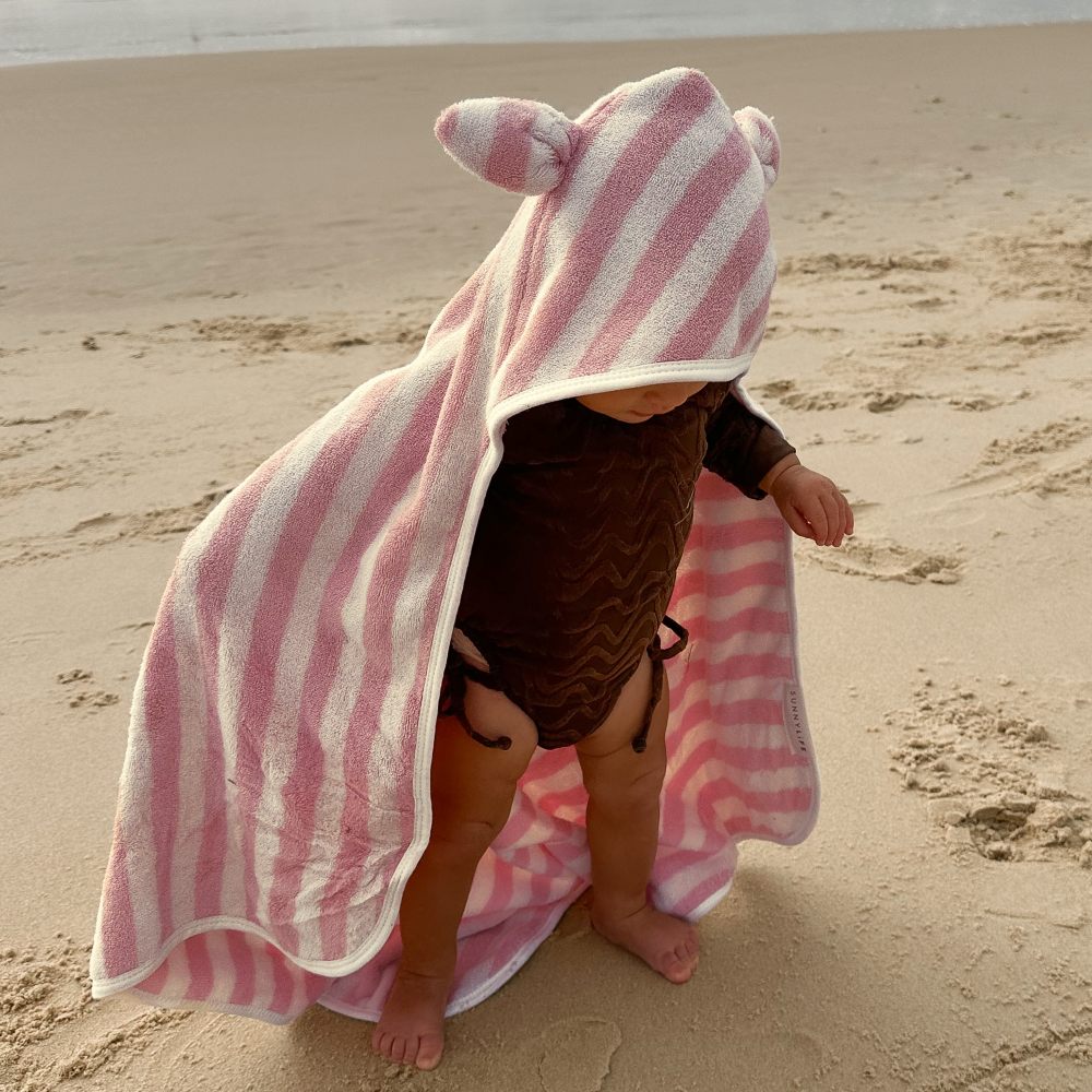 A young child stood on a beach wearing a pink and white hooded towel by SUNNYLiFE