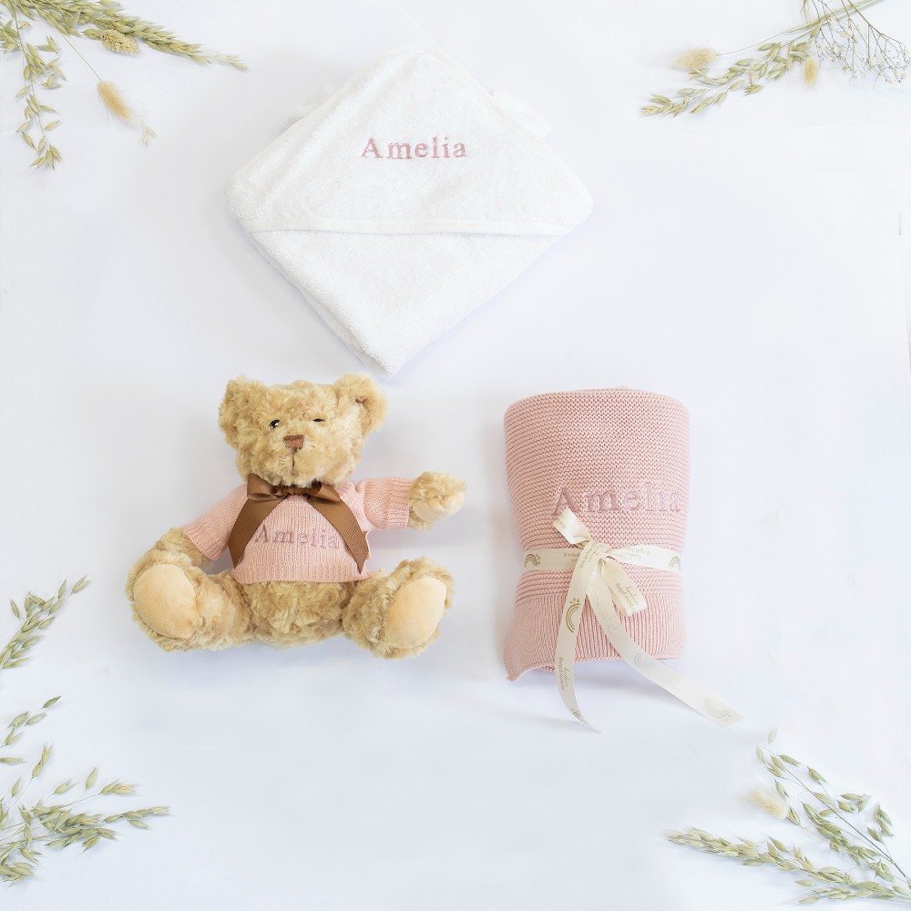 A teddy, pink blanket tied with a satin bow, and a personalised bib by The Baby Gifting Company 