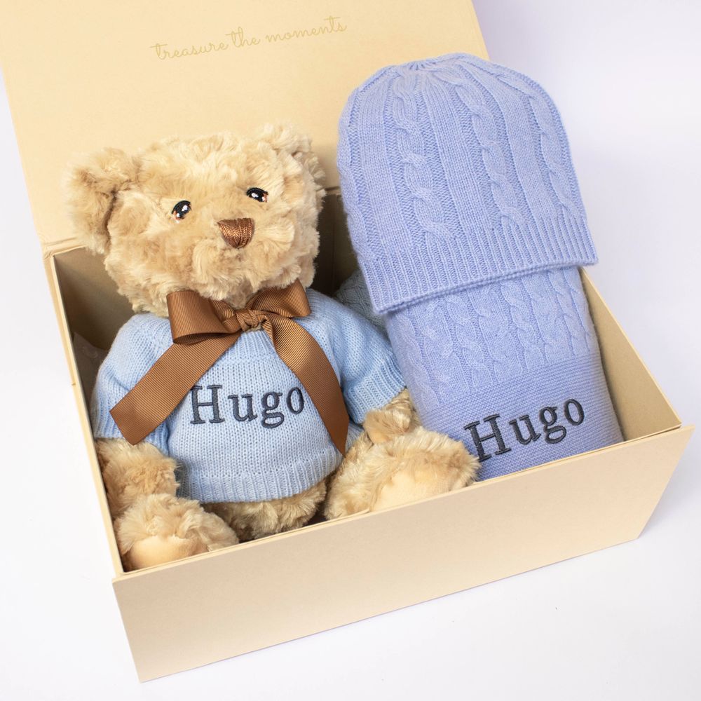 A teddy wearing a blue jumper and a blue blanket displayed in a wooden gift box 