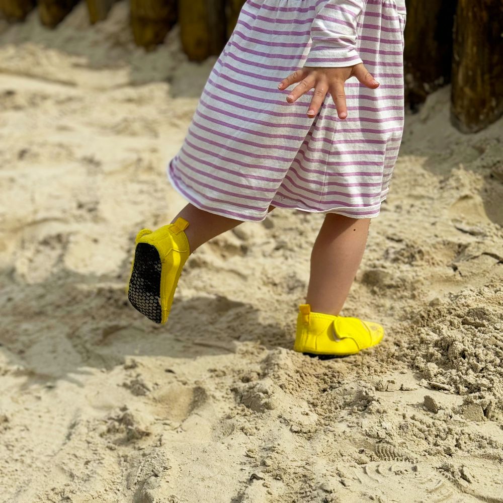 A young girl on a beach wearing a purple and white striped dress and yellow Aqua Socks 
