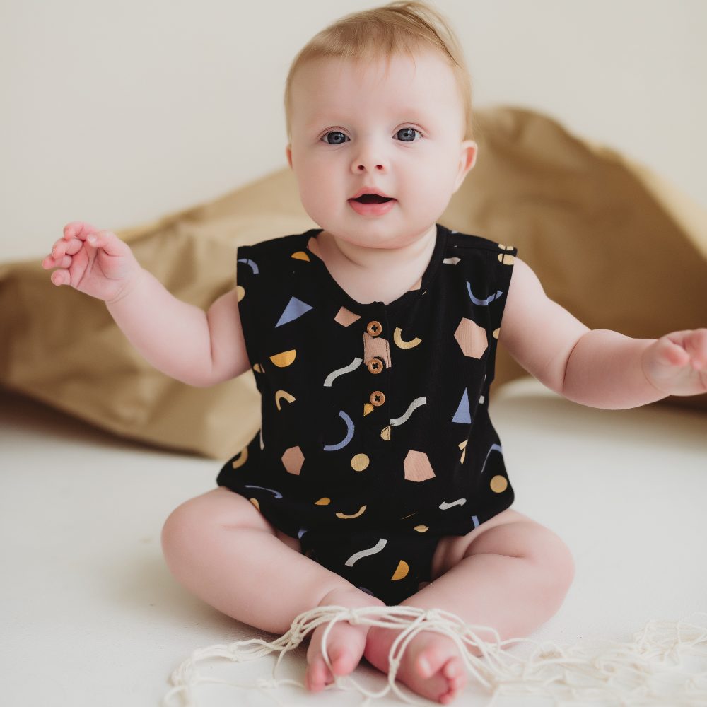 A young baby sat on the floor in front of brown paper wearing a black patterned romper 