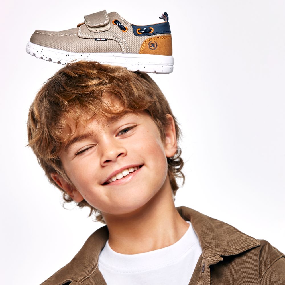 A young smiling boy wearing a white top and brown jacket with a shoe on his head 