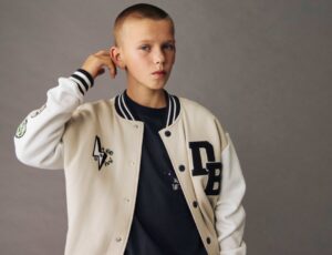 A young boy with his hand raised to his head wearing a Varsity-style jacket from the M&S x The Doodle Boy Collection