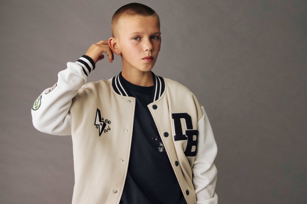 A young boy with his hand raised to his head wearing a Varsity-style jacket from the M&S x The Doodle Boy Collection