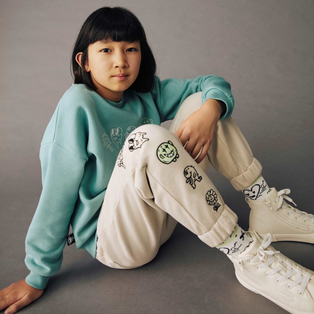 A young girl sat on the floor wearing a blue sweatshirt and cream joggers from the M&S x The Doodle Boy collection 