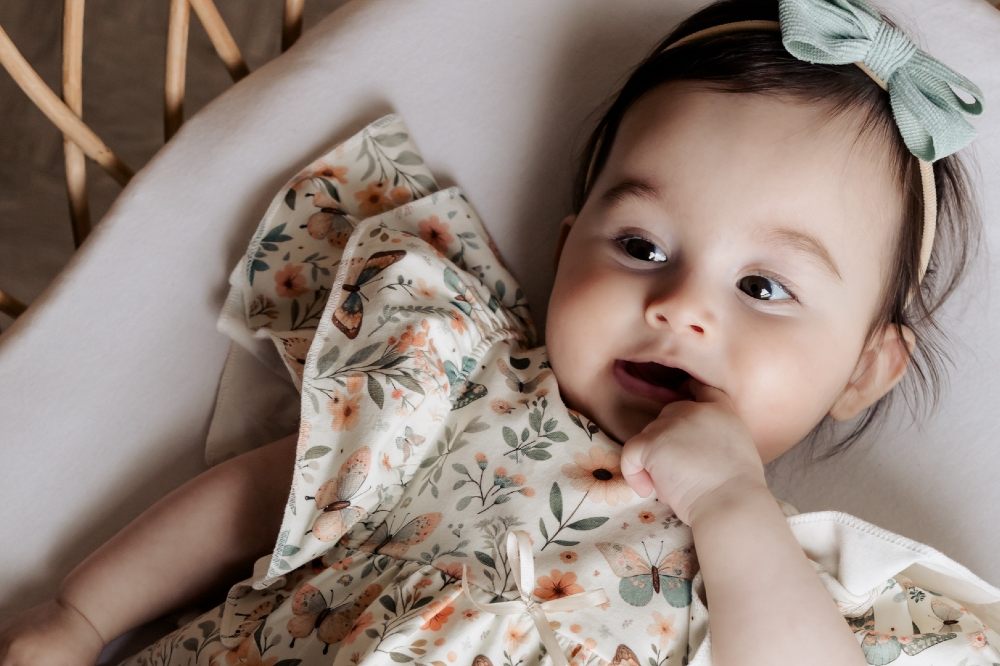 A young baby lying in a cot wearing a floral dress and bow headband 