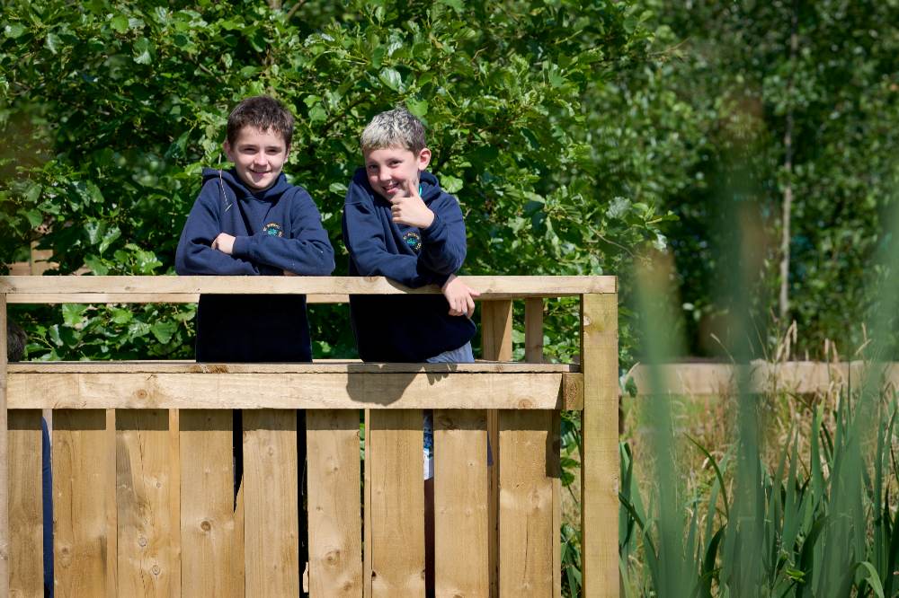 Two boys in school uniform stood on a wooden platform outside 