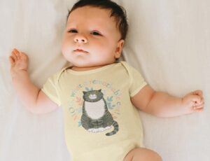 A baby lying on its back wearing a pale yellow babygro with Mog the cat on the front