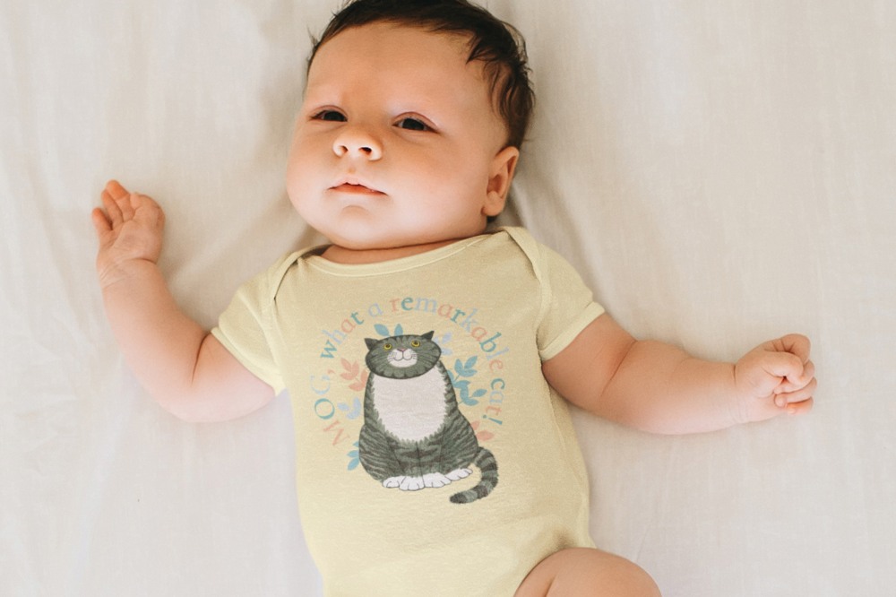 A baby lying on its back wearing a pale yellow babygro with Mog the cat on the front