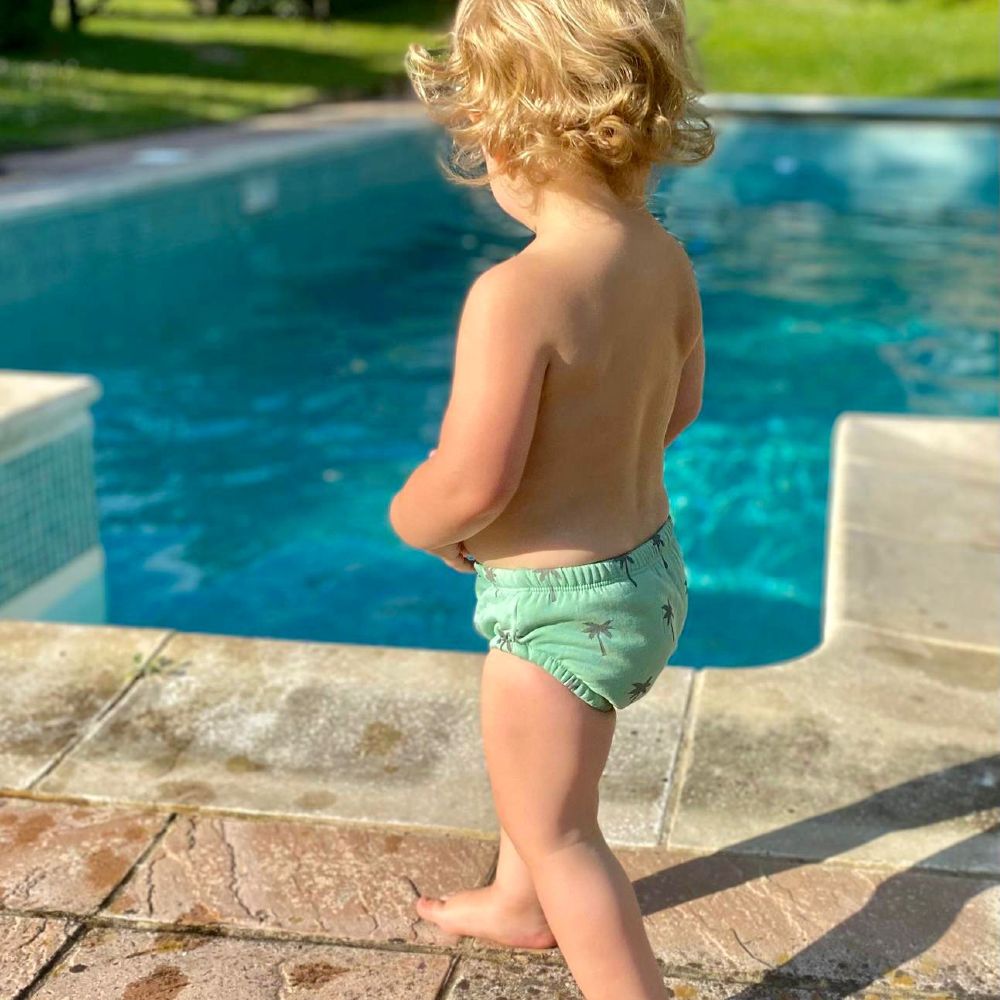 A young child outside wearing a swim nappy stood at the edge of a swimming pool 