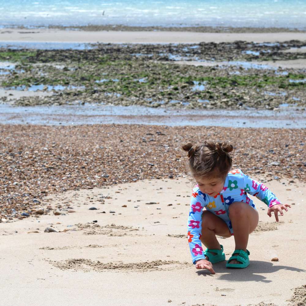 A young girl on a beach in a flower print swimsuit bending down to pick something up