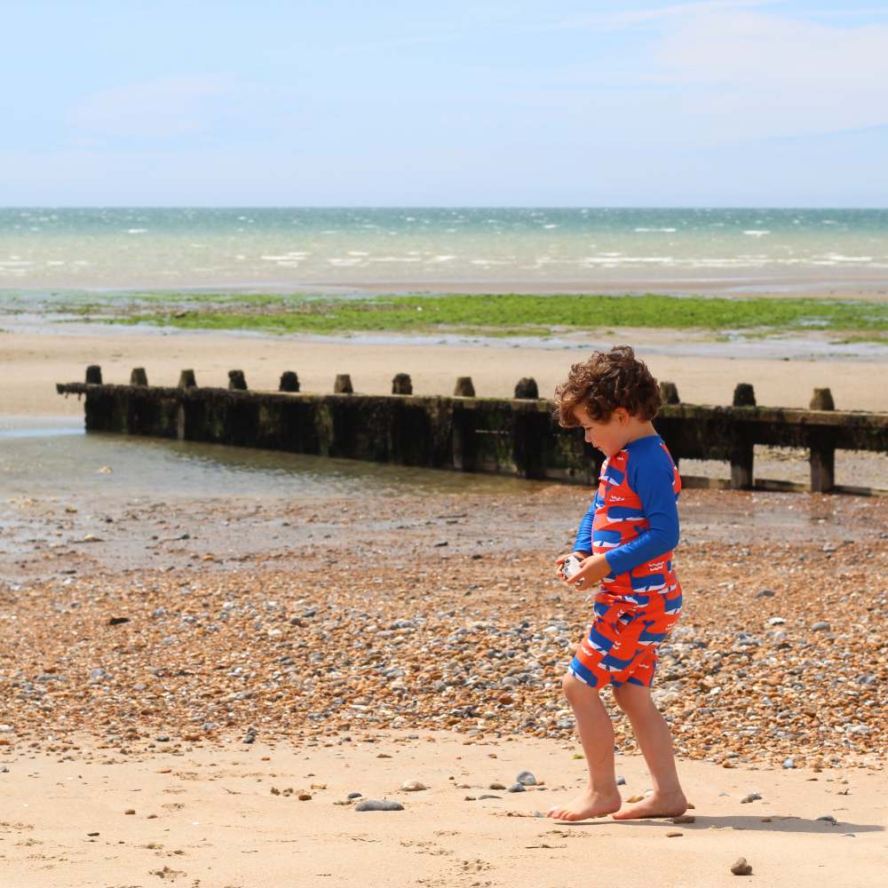 A young boy walking on a beach wearing blue and orange whale print swimwear 