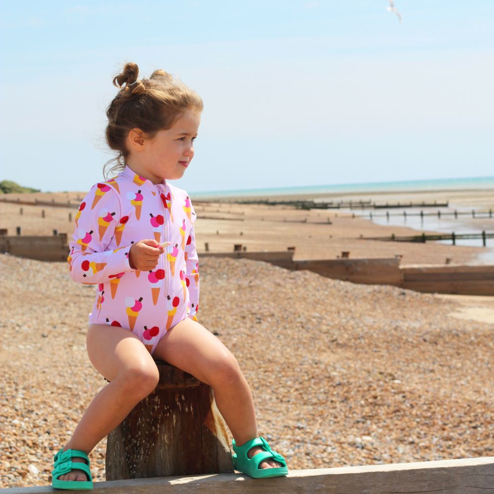 A young girl in an icecream print sunsuit sat on a wooden post on the beach 