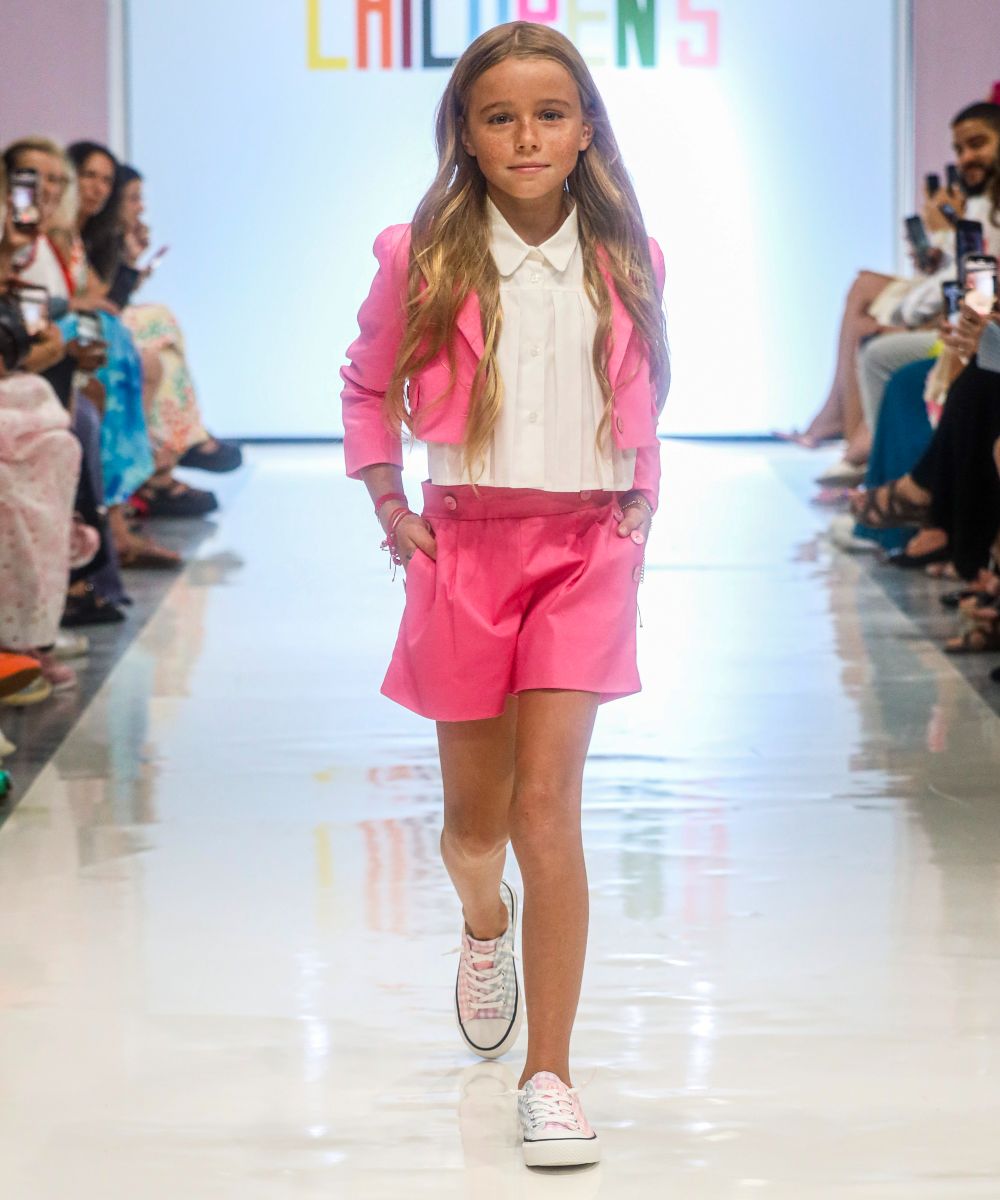 A girl walking on fashion catwalk wearing a pink jacket, white shirt and pink shorts