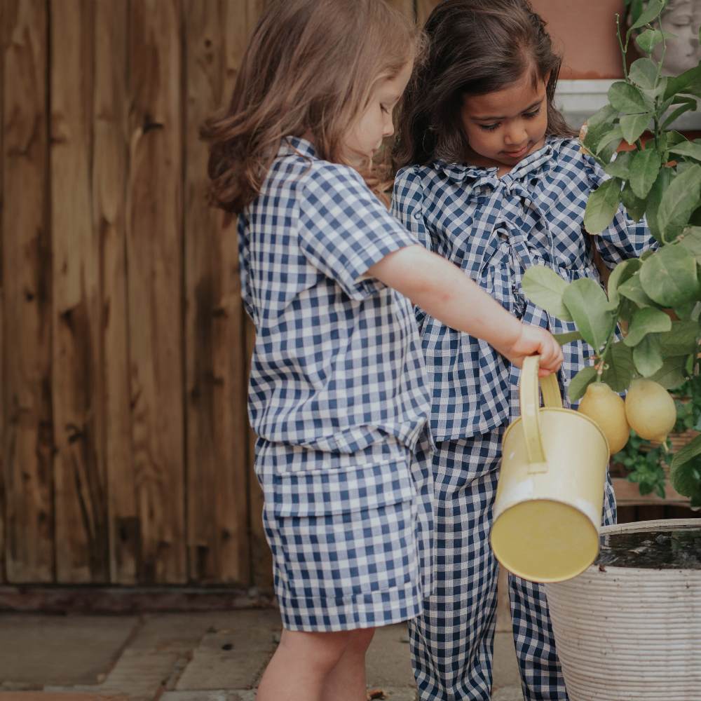 Two children in matching blue and white check outfits outside watering a plant 