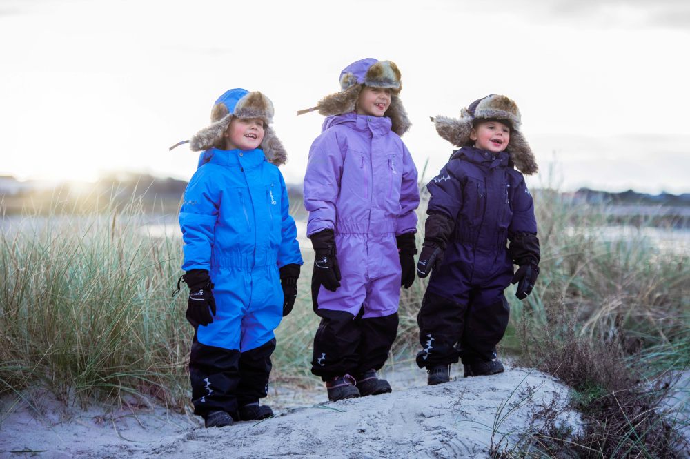 Three children stood outside in snow wearing Penguin Snowsuits by Isbjorn of Sweden