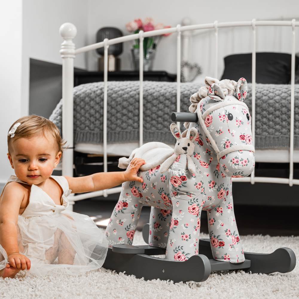 A young girl sat on the floor of a bedroom reaching up to a floral print rocking horse beside her 