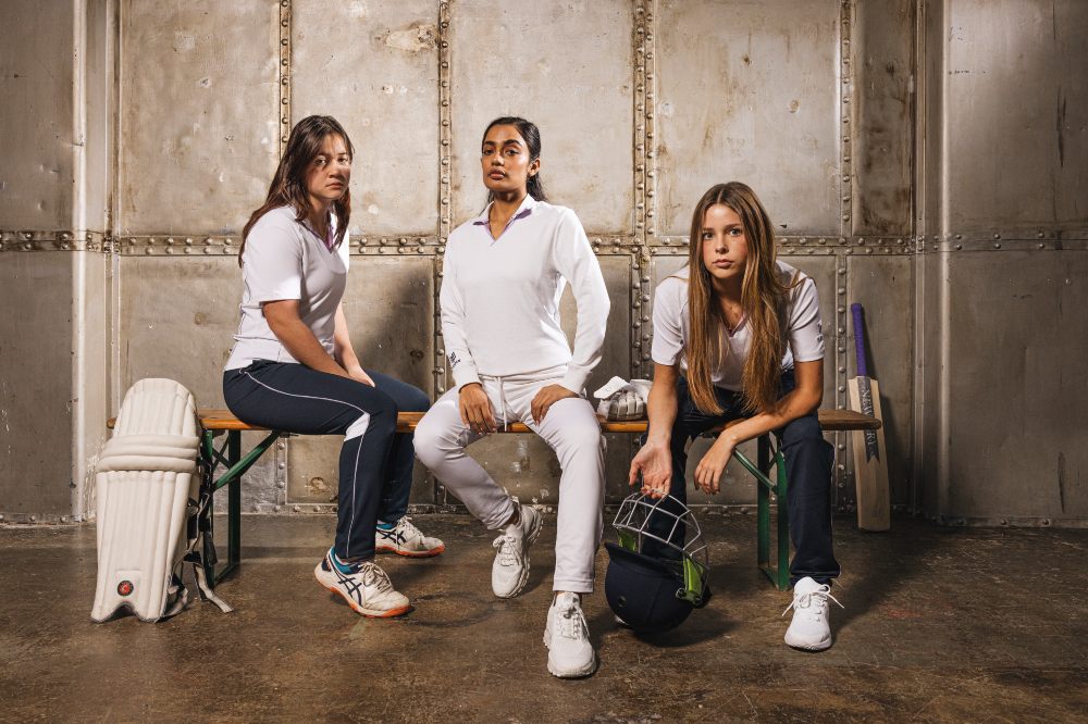 Three girls in cricket clothing sat on a bench in a changing room 