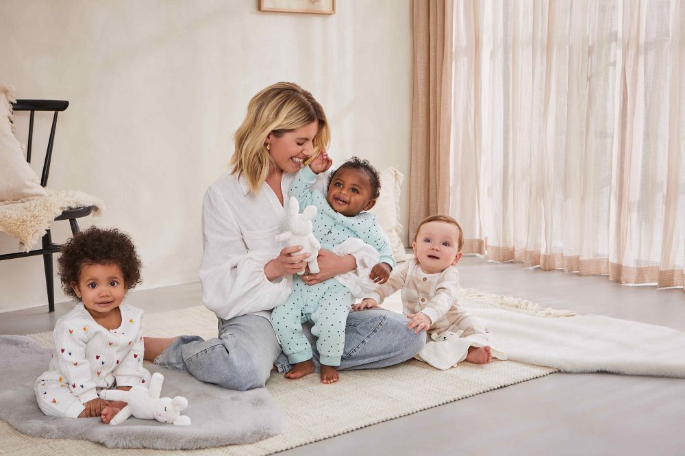 Mollie King sat on the floor with three babies wearing the new Maybe Baby clothing range by Boots