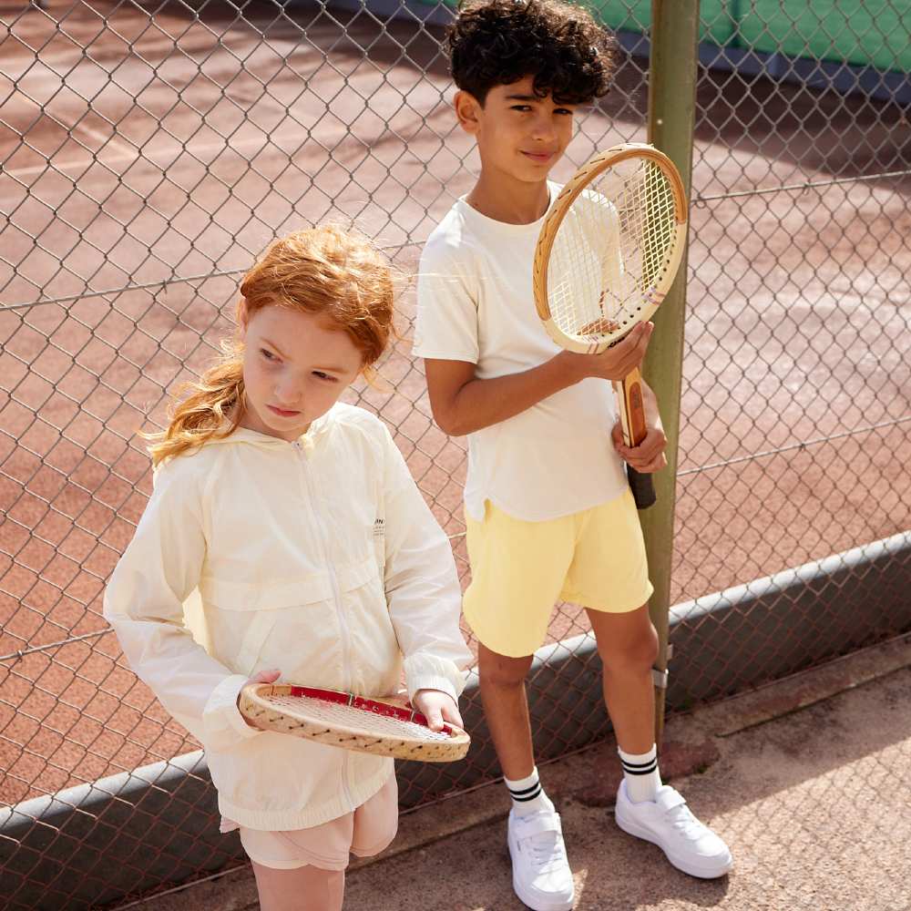 A girl and boy stood on a tennis court holding racquets 