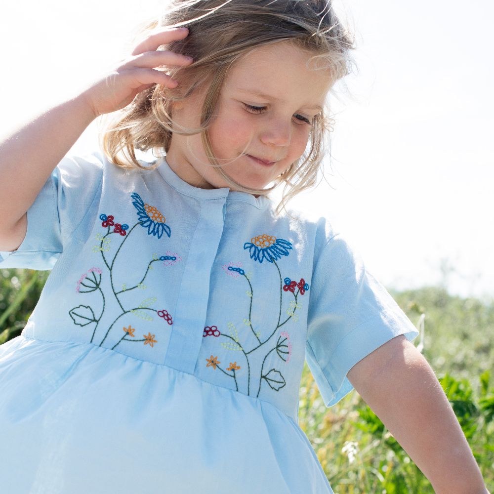 A young girl outside holding back her hair and wearing a blue dress with flower embroidery on the chest