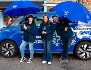 Three people stood with blue umbrellas in front of a blue electric car to promote Small Business Saturday's UK roadshow