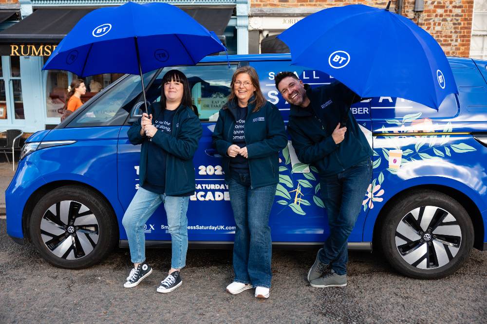 Three people stood with blue umbrellas in front of a blue electric car to promote Small Business Saturday's UK roadshow