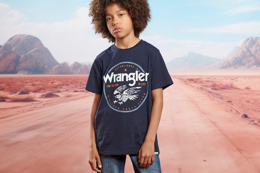 A boy in a Wrangler® T-shirt and jeans stood against a background of a desert road