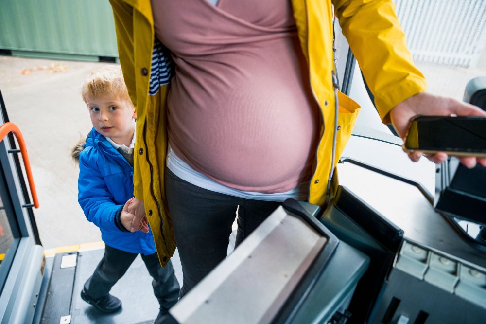 A pregnant woman in a yellow coat getting onto a bus with a young boy in school uniform