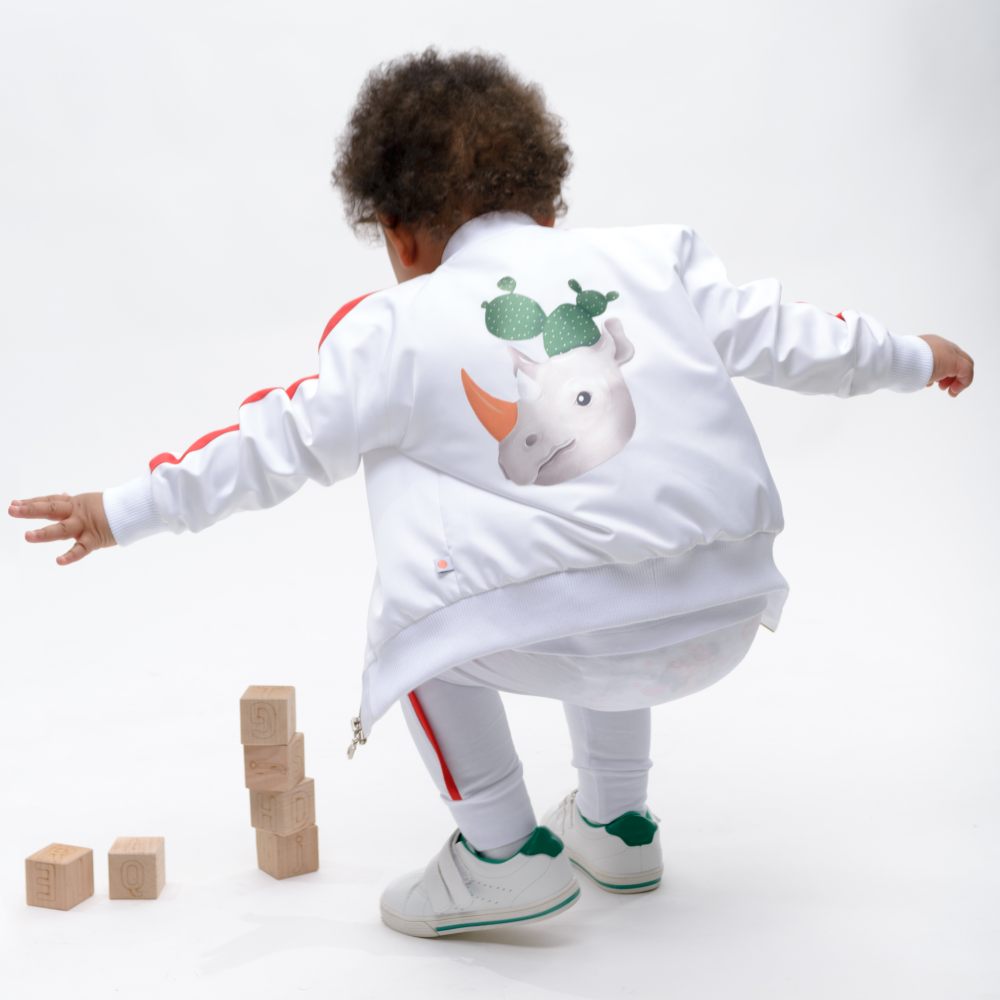 A young child crouching towards some wooden blocks on the floor wearing white trousers and a white jacket with a rhino and cactus illustration on the back by the children's brand ANJOY KIDS