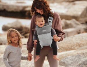 A woman outside holding a young child's hand and carrying a baby on her chest in a BabyBjörn carrier