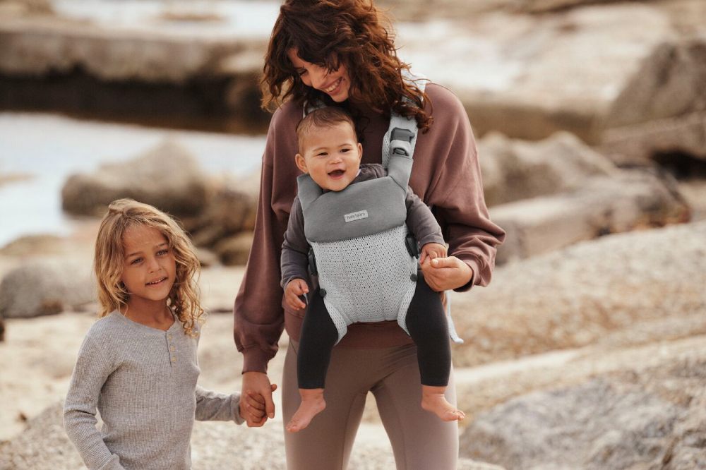 A woman outside holding a young child's hand and carrying a baby on her chest in a BabyBjörn carrier