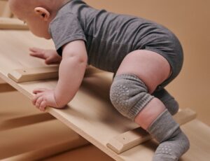 A baby in a grey babygro, kneepads and socks climbing up a wooden frame