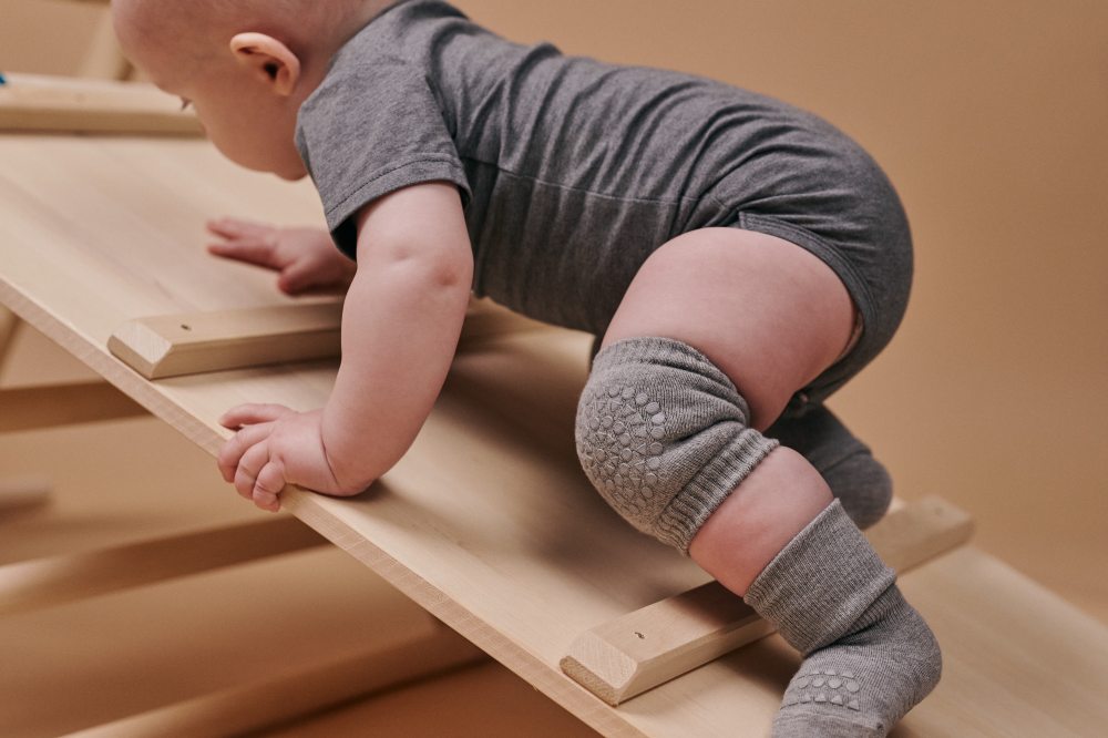 A baby in a grey babygro, kneepads and socks climbing up a wooden frame