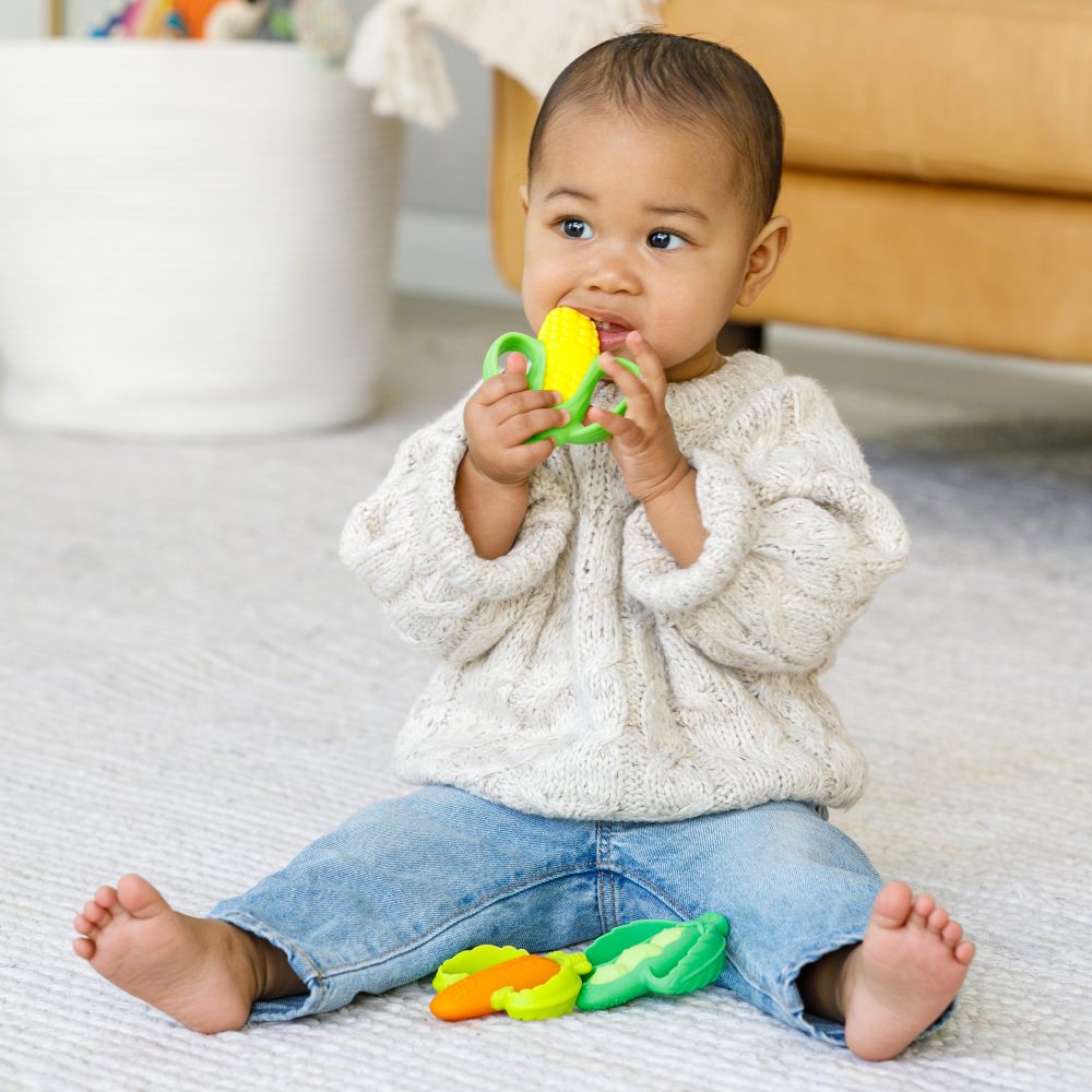 A baby sat on the floor with three vegetable shaped teethers 